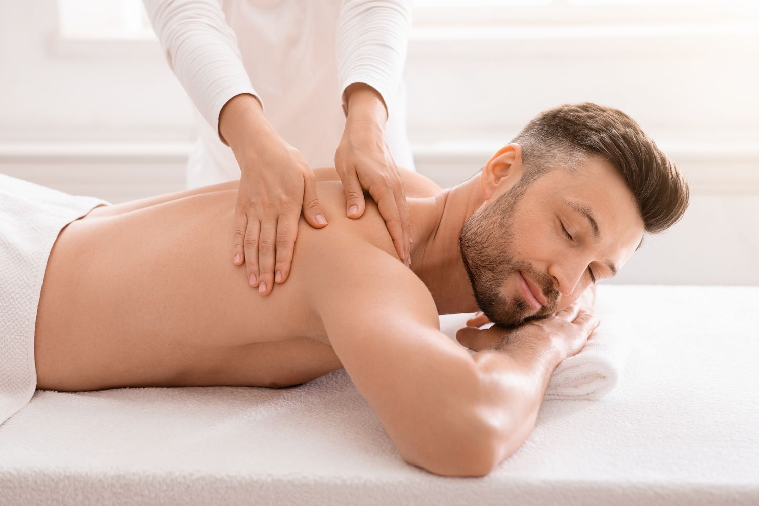 Unrecognizable female therapist rubbing joyful man shoulders at modern spa salon, side view. Handsome bearded middle-aged man relaxing while getting healing back massage